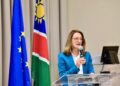 BEATRIZ MARTINS1 - The Brief | Namibia's Leading Business & Financial News Woman in a blue blazer speaks into a handheld microphone at a clear podium, with European and other flags in the background.