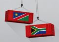 Nam X SA - The Brief | Namibia's Leading Business & Financial News Two red shipping containers bearing the flags of Namibia and South Africa being hoisted by cranes against a gray sky.