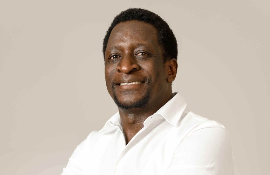 Amos Shiyuka - The Brief | Namibia's Leading Business & Financial News Portrait of a smiling Black man in a white button-down shirt against a neutral beige background, studio lighting.