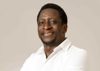 Portrait of a smiling Black man in a white button-down shirt against a neutral beige background, studio lighting.
