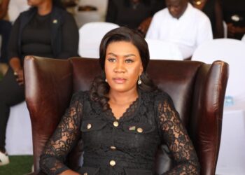 Woman in a black lace dress sits in a brown leather high-back chair at a formal event, with attendees behind her.