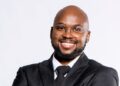 Nesdha De Jongh - The Brief | Namibia's Leading Business & Financial News Smiling professional portrait: a man in a black suit and tie with round glasses, facing the camera.
