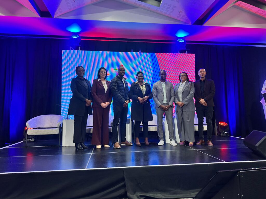 WhatsApp Image 2026-07-08 at 20307 PM - The Brief | Namibia's Leading Business & Financial News Eight diverse professionals in business attire stand together on a stage with a colorful LED backdrop and purple-blue lighting, posing for a group photo at an event.