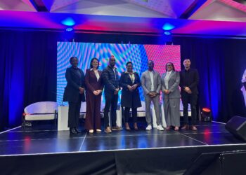 Eight diverse professionals in business attire stand together on a stage with a colorful LED backdrop and purple-blue lighting, posing for a group photo at an event.