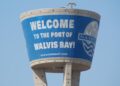 Blue water-tole sign on a cylindrical water tower that reads 'Welcome to the Port of Walvis Bay!' against a clear sky with a port logo on the right side.