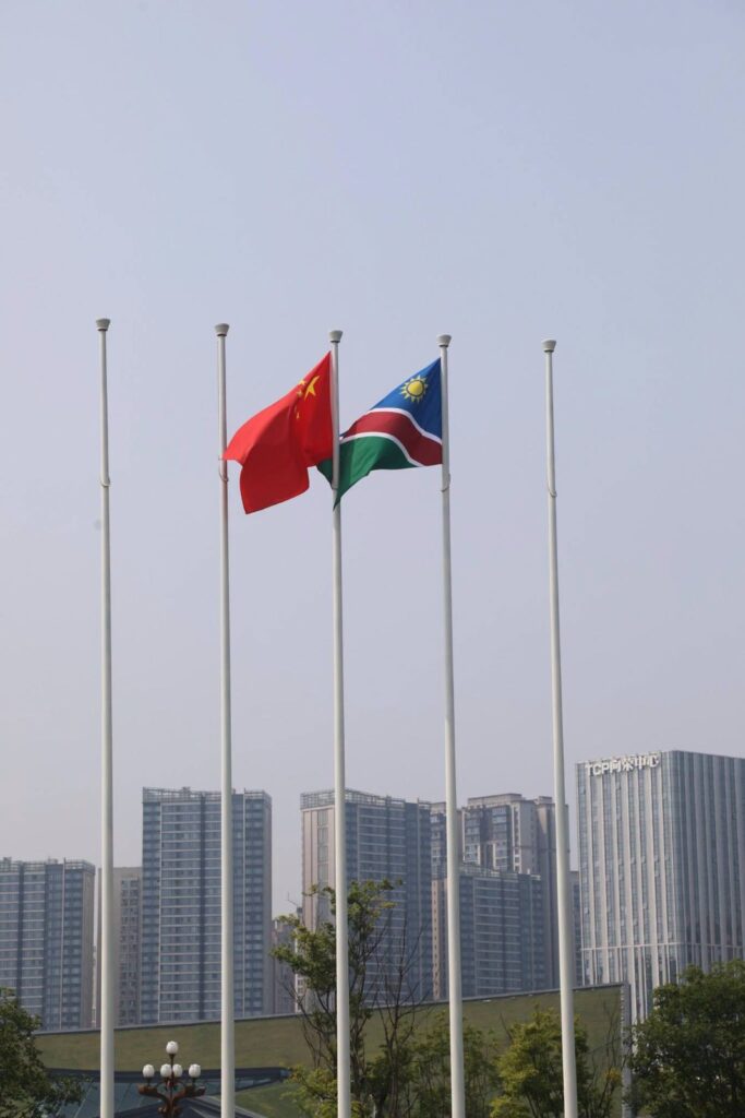 China and Namibia national flags flying on flagpoles above a city skyline in the background.