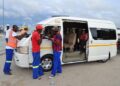 PUBLIC TRANSPORT - The Brief | Namibia's Leading Business & Financial News White minivan with a yellow stripe parked on a dirt road; volunteers in safety vests and casual clothes assist passengers at the open door.