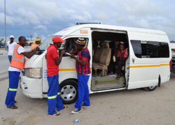 White minivan with a yellow stripe parked on a dirt road; volunteers in safety vests and casual clothes assist passengers at the open door.
