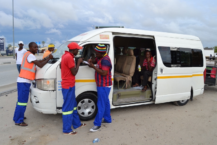 PUBLIC TRANSPORT - The Brief | Namibia's Leading Business & Financial News White minivan with a yellow stripe parked on a dirt road; volunteers in safety vests and casual clothes assist passengers at the open door.