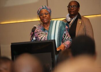 Older woman wearing a colorful headwrap and traditional attire speaks at a podium with a microphone, while another woman stands behind her in a blazer.