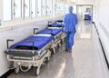 Hospital hallway with a row of patient stretchers covered in blue sheets along the left wall; a medical professional in blue scrubs walks away down the corridor