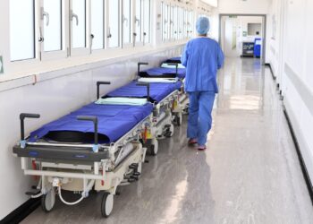 Hospital hallway with a row of patient stretchers covered in blue sheets along the left wall; a medical professional in blue scrubs walks away down the corridor