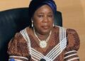 Portrait of an African woman wearing a blue headwrap and a brown patterned dress with white trim, seated at a desk with a microphone in front, suggesting a press conference.