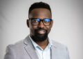 Professional headshot of a smiling Black man wearing glasses and a light gray blazer against a neutral backdrop.