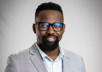 Professional headshot of a smiling Black man wearing glasses and a light gray blazer against a neutral backdrop.