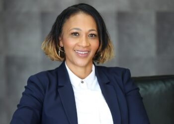 Professional head-and-shoulders portrait of a smiling woman in a navy blazer and white shirt, with gold hoop earrings, against a gray textured background.