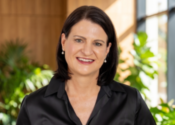 Smiling woman in a black blouse stands in a bright, plant-filled office background.