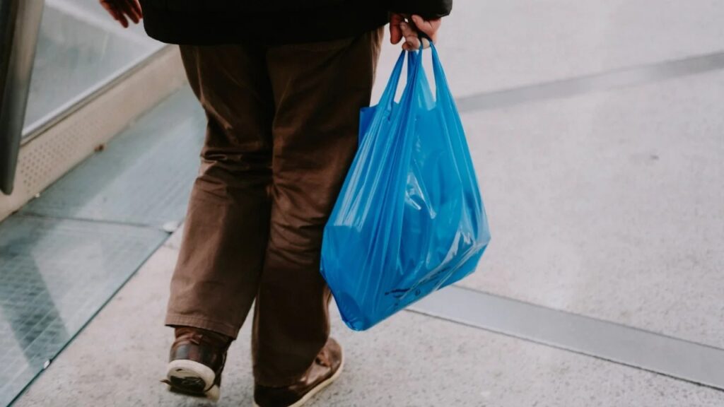 Person walking on an urban sidewalk, holding a bright blue plastic shopping bag.