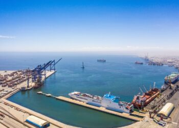 Industrial port with container cranes and a white passenger ship docked at a quay, blue sea and clear sky.