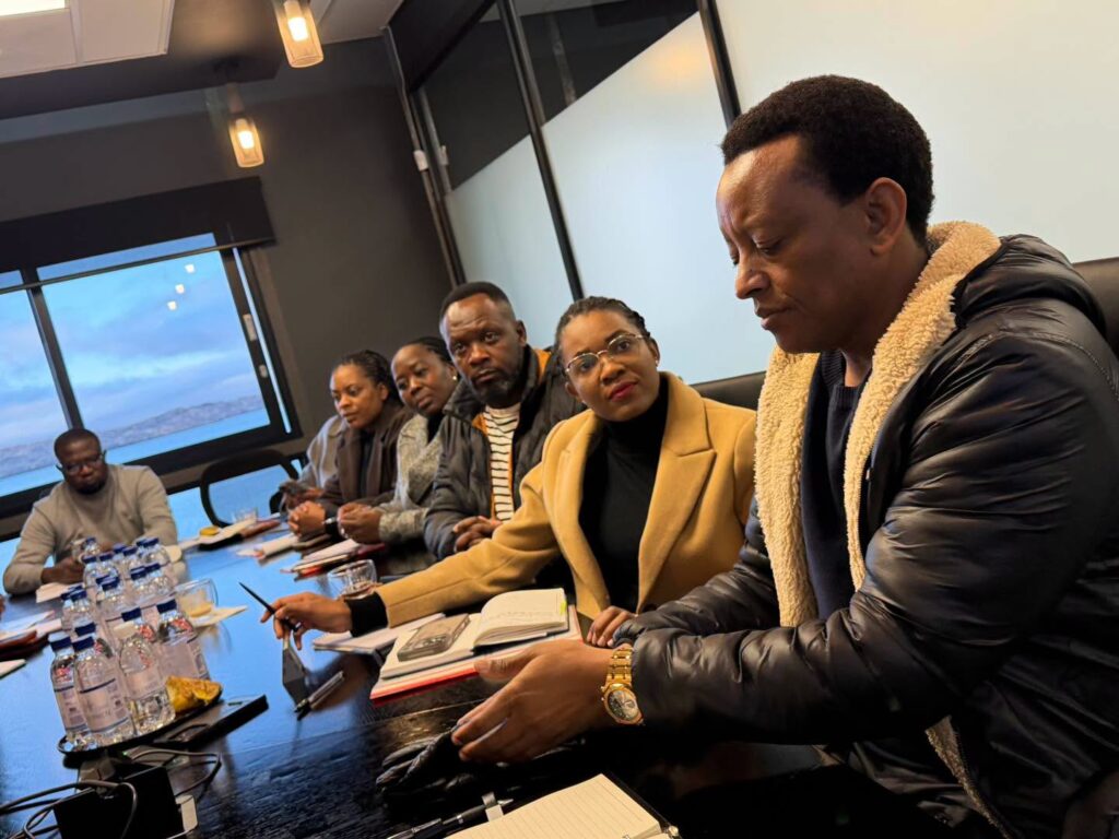 Seven people sit around a conference table in a meeting, with one man in a black shearling jacket closest to the camera.