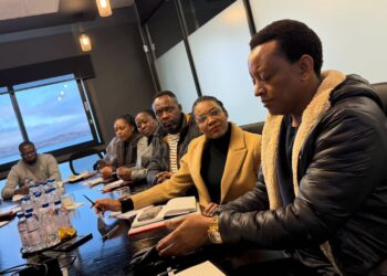 Seven people sit around a conference table in a meeting, with one man in a black shearling jacket closest to the camera.