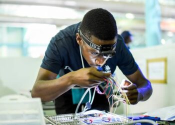 Young person wearing a headlamp soldering colorful wires at a lab workbench in a bright workshop.
