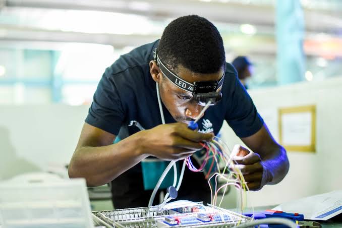 Young person wearing a headlamp soldering colorful wires at a lab workbench in a bright workshop.