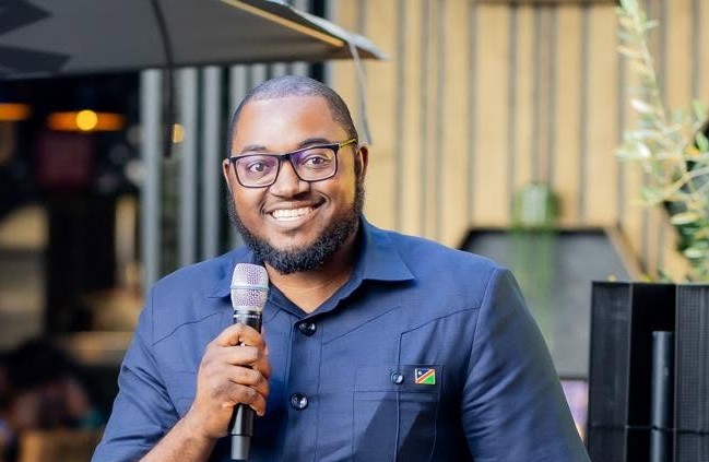 NUJOMA JUNIOR - The Brief | Namibia's Leading Business & Financial News Smiling man wearing glasses and a blue shirt, holding a microphone while speaking at an outdoor event.