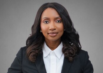 Professional woman in a black blazer and white shirt, facing the camera against a gray backdrop (head-and-shoulders portrait).