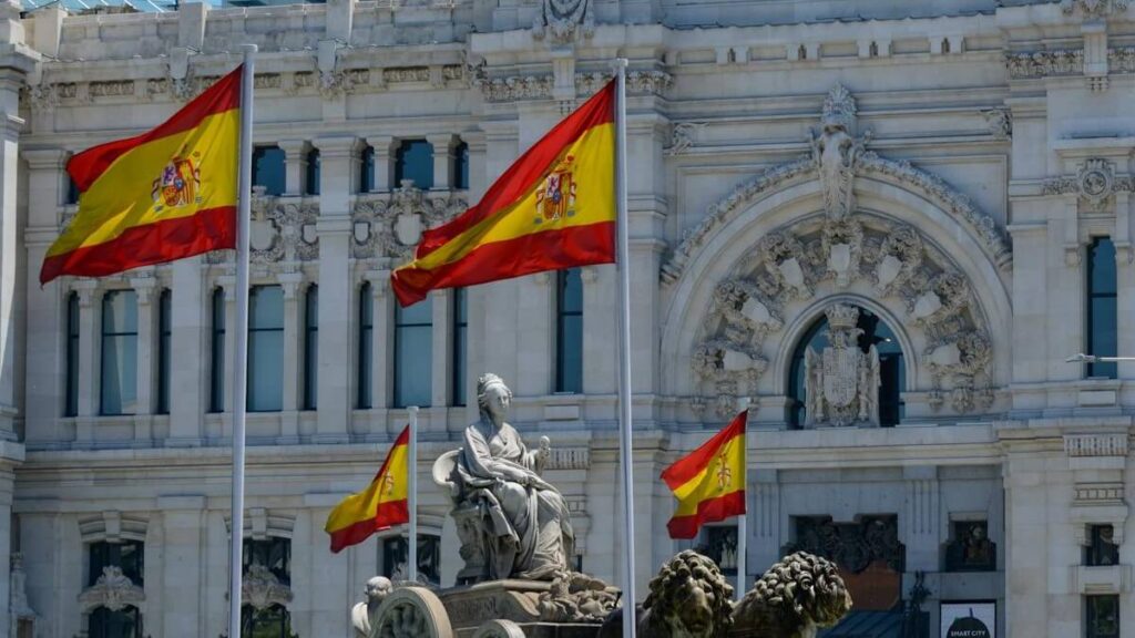 SPAIN FLAG - The Brief | Namibia's Leading Business & Financial News Spanish flags fly in front of a grand white stone building with intricate carvings and arched entrance, and a seated statue fountain in the foreground.
