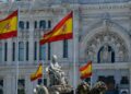 Spanish flags fly in front of a grand white stone building with intricate carvings and arched entrance, and a seated statue fountain in the foreground.