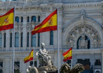 Spanish flags fly in front of a grand white stone building with intricate carvings and arched entrance, and a seated statue fountain in the foreground.