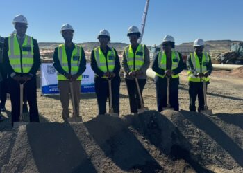 Six construction workers wearing hard hats and neon vests stand with shovels in front of a dirt mound at a groundbreaking event on an arid construction site.