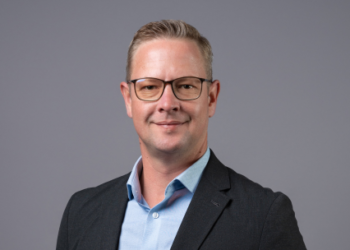 Professional headshot: man in a dark blazer and light blue shirt, smiling at the camera against a gray backdrop.