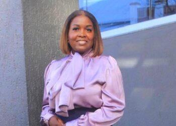 Woman with shoulder-length hair wearing a lavender blouse with a large bow, smiling outdoors beside a textured wall and glass window.