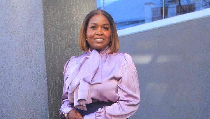 Woman with shoulder-length hair wearing a lavender blouse with a large bow, smiling outdoors beside a textured wall and glass window.