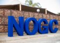 Large blue 3D letters spelling a word along a sidewalk, with a gabion wall behind and palm trees in the background.