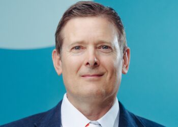 Head-and-shoulders portrait of a smiling man in a navy suit and white shirt against a teal background.