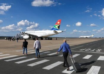 Passengers walk on the tarmac toward a South African Airways plane parked at a gate, under a blue sky with white clouds.