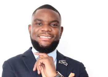Professional headshot of a smiling Black man in a navy suit, light blue shirt, orange tie, and pocket square, looking at the camera.