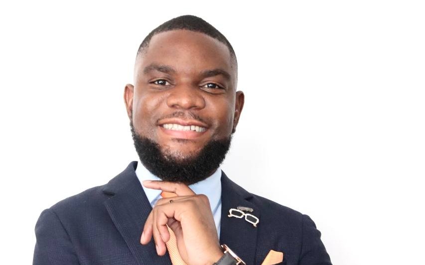 Professional headshot of a smiling Black man in a navy suit, light blue shirt, orange tie, and pocket square, looking at the camera.