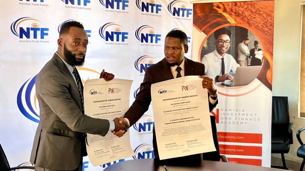 Two suited men shake hands and display signed memorandum papers at a Namibia Trade Forum backdrop.