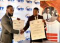 Two suited men shake hands and display signed memorandum papers at a Namibia Trade Forum backdrop.