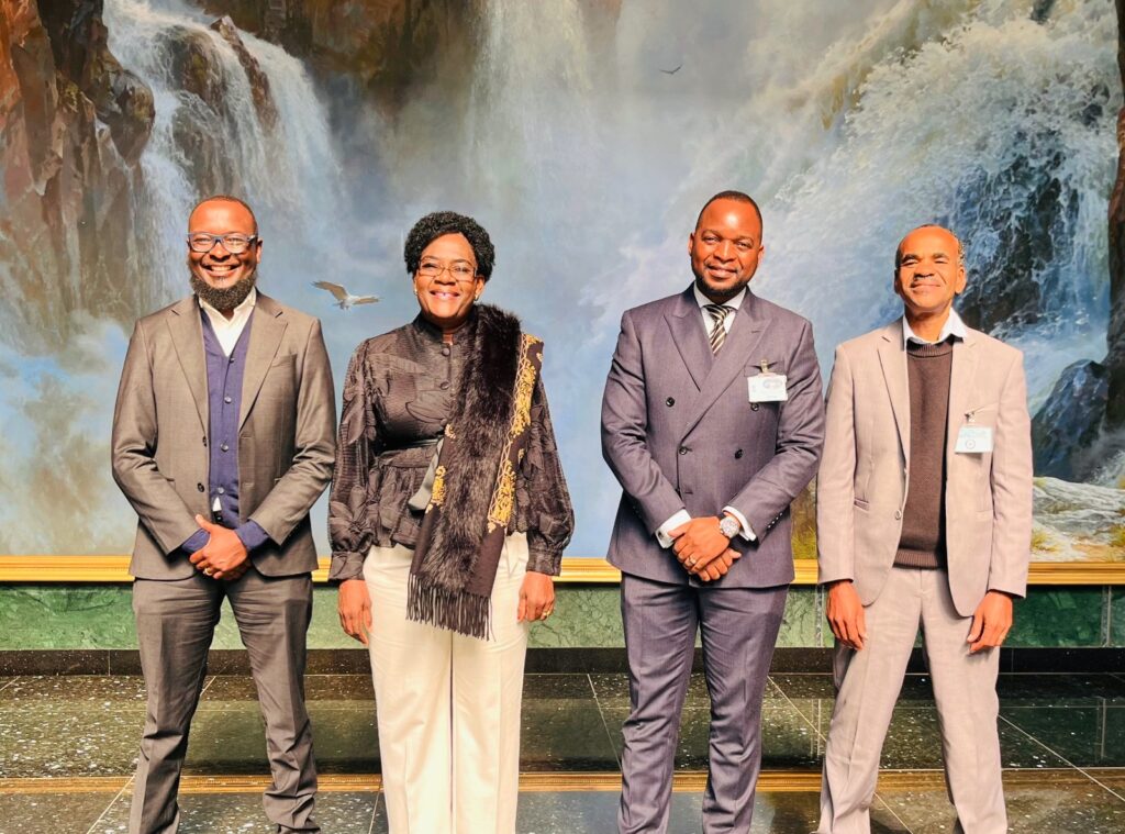 WhatsApp Image 2026-07-21 at 43514 PM - The Brief | Namibia's Leading Business & Financial News Four professionally dressed individuals pose together for a group photo in front of a waterfall backdrop, smiling at the camera in a formal setting.