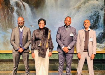 Four professionally dressed individuals pose together for a group photo in front of a waterfall backdrop, smiling at the camera in a formal setting.