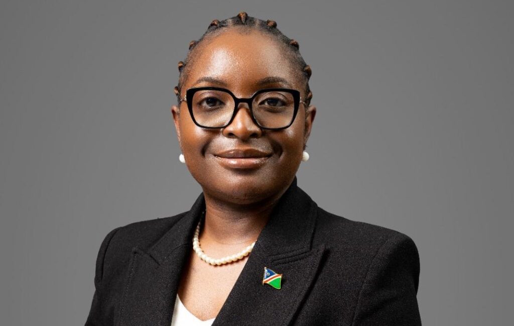 Professional head-and-shoulders portrait of a Black woman in a blazer and pearls, wearing glasses with a flag pin on her lapel.