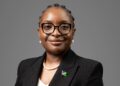 Professional head-and-shoulders portrait of a Black woman in a blazer and pearls, wearing glasses with a flag pin on her lapel.