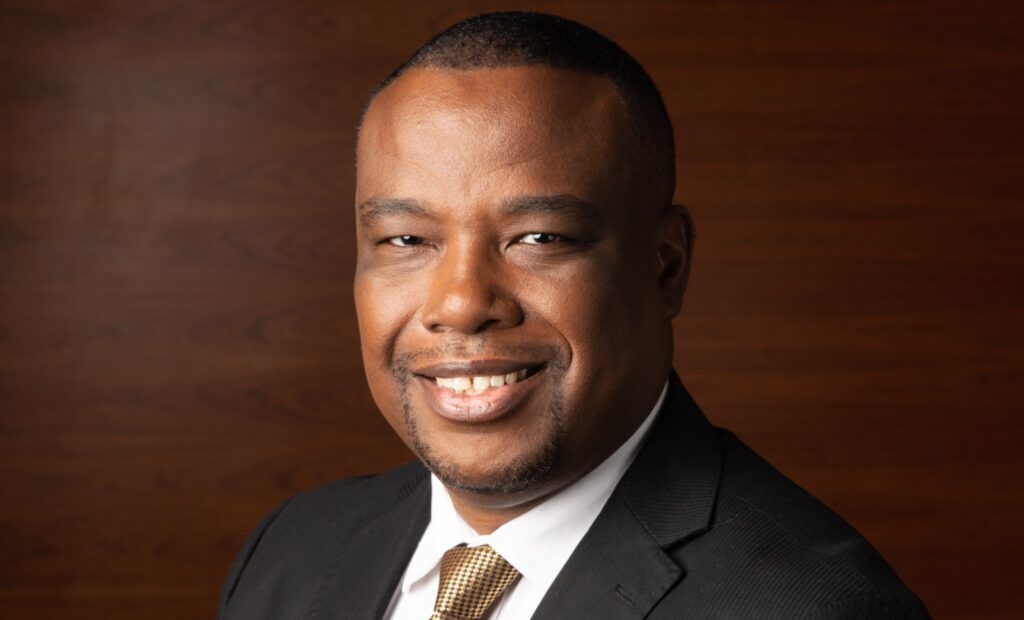Erich Gariseb - The Brief | Namibia's Leading Business & Financial News Smiling man in a black suit with a gold tie, head-and-shoulders portrait against a wooden backdrop.