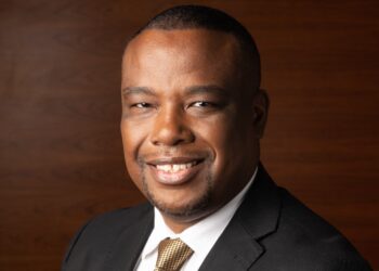Smiling man in a black suit with a gold tie, head-and-shoulders portrait against a wooden backdrop.
