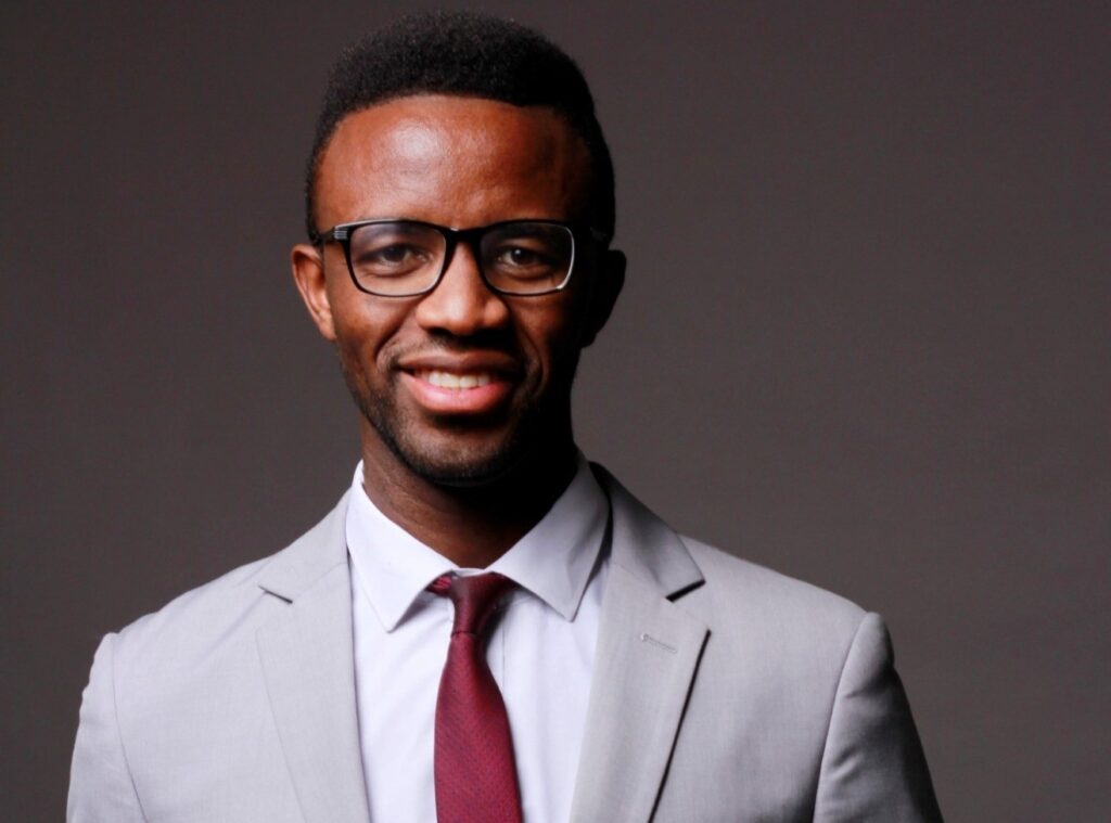 Professional headshot of a smiling man in a light gray suit, white shirt, burgundy tie, and glasses against a dark backdrop.